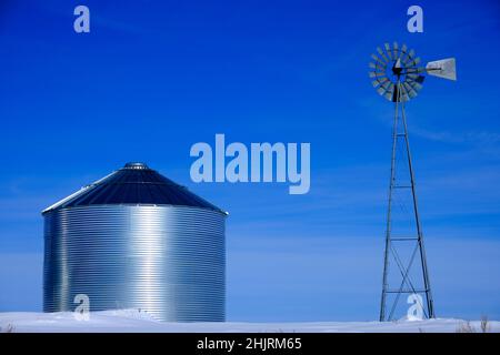 Windmühle und Stahlkornsilos im Winterschnee mit blauem Himmel auf dem Bauernhof für die landwirtschaftliche Landwirtschaft Stockfoto