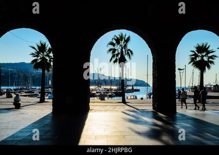 Schöner Blick auf die Marina, das Meer und die Boote durch ein antikes Bogengebäude in Koper, Slowenien. Silhouetten von Palmen und Menschen an einem sonnigen Tag Stockfoto