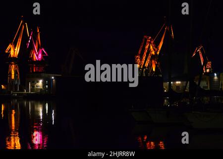 Große beleuchtete Krane auf der Werft Pula in Kroatien bei Nacht. Die Farben spiegeln sich im Dunklen Wasser des Hafens wider. LED-Licht-Choreographie. Stockfoto