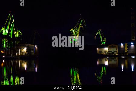 Große beleuchtete Krane auf der Werft Pula in Kroatien bei Nacht. Grüne Farben spiegeln sich im Dunklen Wasser des Hafens wider. LED-Licht-Choreographie. Stockfoto