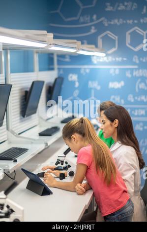 Mädchen in der Nähe des Mikroskops und beobachtende Lehrerin Stockfoto