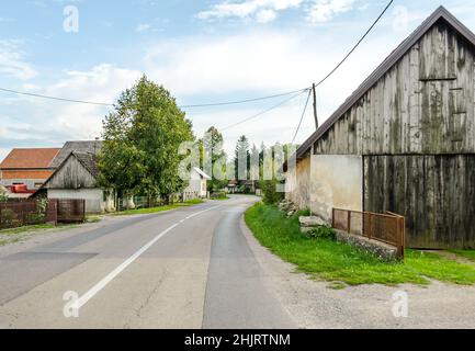 Fahren auf einer Provinzstraße durch Dörfer in der kroatischen Landschaft. Ländliche Landschaft mit grünen Bäumen und alten Hütten an einem sonnigen Tag. Stockfoto