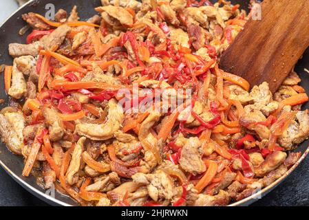 Ein Holzspatel mischt Schweinefleisch in einer Pfanne mit Paprika, Karotten, Zwiebeln. Nahaufnahme. Stockfoto