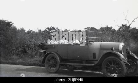 Um 1920s, historisch, auf einer Landstraße, eine Familie...Vater, Mutter, Schwester und kleines Kind, das in einem offenen Wagen der damaligen Zeit saß, England, Großbritannien. Alle drei Erwachsenen tragen Hüte, wie sie es in dieser Zeit taten. An der Fahrertür ragt von unten ein Anlassergriff heraus und die Hupe ist eine einfache Gummihupe, die neben dem Lenkrad montiert ist. Stockfoto