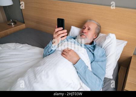 High-Angle-Seitenansicht des gebärteten älteren Mannes, der sich einen Film ansieht, der im Schlafzimmer zu Hause auf dem Bett liegt. Stockfoto