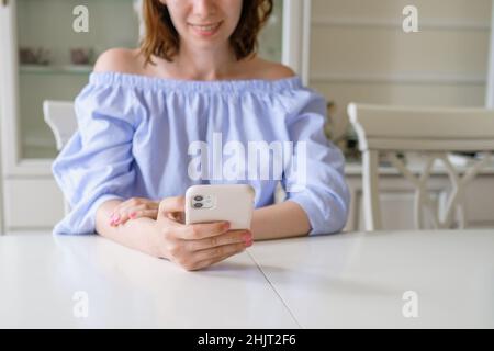 Junge glückliche Brünette Frau in stilvoller Bluse prüft Mail im Handy sitzen am weißen Tisch in den frühen Morgen zu Hause aus der Nähe Stockfoto