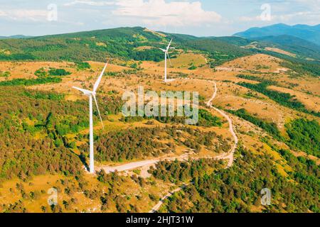 Windmühlen mit großen Klingen werden in Waldbergen betrieben und erzeugen unter bewölktem Himmel am Sommertag saubere Energie von der Drohne aus Stockfoto