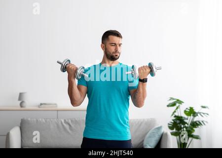 Seriöser, tausendjähriger europäischer Athlet in blauem T-Shirt führt Kraftübungen für Arme und Hanteln durch Stockfoto