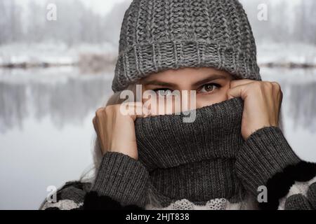 Portrait einer Frau im Winter in Strickmütze und Pullover. Das Mädchen bedeckt die Hälfte ihres Gesichts. Stockfoto