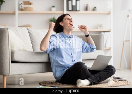 Überglücklich freiberuflicher Mann, der mit einem Laptop den Erfolg emotional feiert, auf dem Boden sitzt und die Hände vor Freude hebt Stockfoto