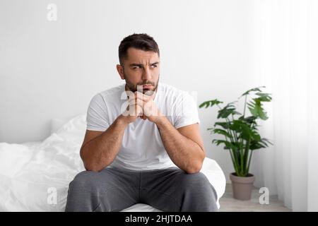 Unglückliche Depression europäischer Millennials-Mann in weißem T-Shirt leidet an Problemen sitzt auf dem Bett im Schlafzimmer Stockfoto