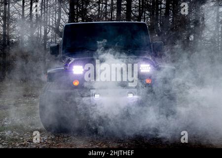 Schwarzer Jeep-Wrangler auf einer unbefestigten Straße mit Scheinwerfern und Rauch vor und Bäumen im Hintergrund für das ultimative Offroad-Erlebnis Stockfoto