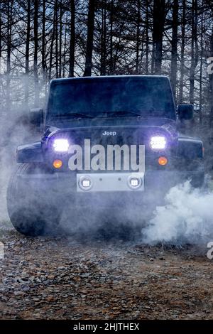 Schwarzer Jeep-Wrangler auf einer unbefestigten Straße mit Scheinwerfern und Rauch vor und Bäumen im Hintergrund für das ultimative Offroad-Erlebnis Stockfoto