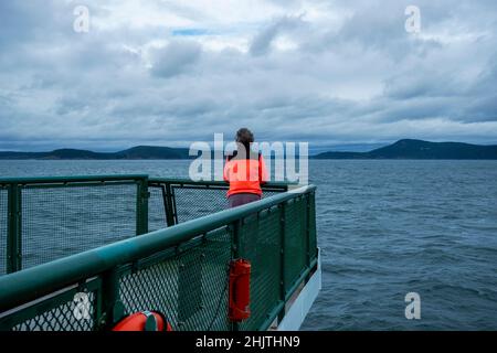 Blick auf eine ältere Frau in einer hellen, orangefarbenen Jacke, die von der Washington State Ferry nach San Juan Island auf den Pazifik blickt Stockfoto