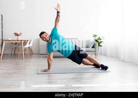 Lächelnder, fröhlicher, tausendjähriger europäischer Athlet, der im minimalistischen Wohnzimmer an der Matte eine Seitenwand macht Stockfoto