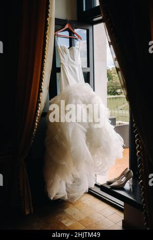 Ein weißes Hochzeitskleid hängt an einem Kleiderbügel in der Nähe des Fensters. Stockfoto