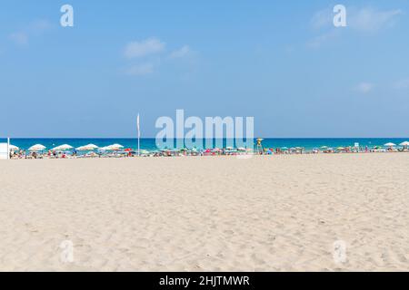 Großer Strand mit Badegästen am Ufer, die sich unter farbigen Sonnenschirmen vor den Sonnenstrahlen schützen. Menschen im Bild sind nicht erkennbar. Stockfoto