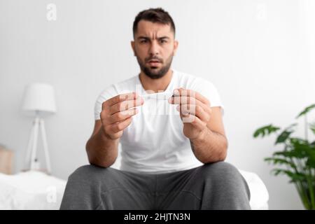 Trauriger junger Mann aus europa im weißen T-Shirt leidet unter gesundheitlichen Problemen, schaut auf Thermometer sitzt auf dem Bett Stockfoto