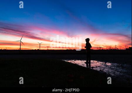 Silhouette eines Jungen, der während des Sonnenuntergangs in einer Pfütze auf einem Windpark steht Stockfoto