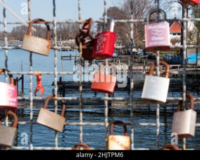 Liebesschlösser hängen an einem Gitter, im Hintergrund ist ein See mit einem Yachthafen Stockfoto