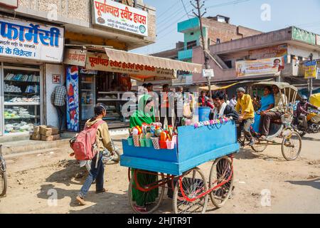 Geschäftige Straßenszene mit Geschäften am Straßenrand und einem Schubkarren, der Haushaltswaren verkauft, am Stadtrand von Varanasi (ehemals Benares), Uttar Pradesh, Nordindien Stockfoto