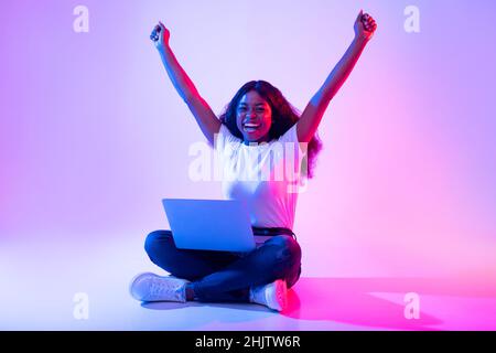 Überglücklich junge schwarze Hündin mit Laptop, der die Arme hochhebt und Erfolge im Neonlicht feiert Stockfoto