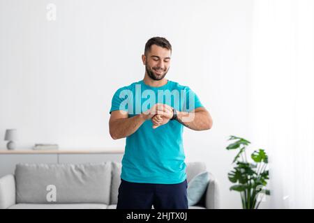 Froh glücklich Millennial europäischen Kerl Athlet in Uniform Kontrolle Puls auf Fitness-Tracker im Wohnzimmer Innenraum Stockfoto