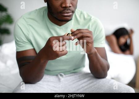 Verärgert traurig junge schwarze Mann sitzt auf dem Bett und zieht aus Ehering, ignoriert beleidigte Frau nach Streit Stockfoto