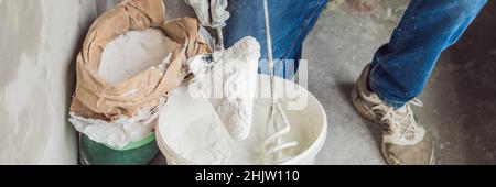 Der junge Maler knetet Kitt mit Wasser in einem Eimer mit einem Handmixer für das Bauen von MISCHBANNERN im langen Format Stockfoto