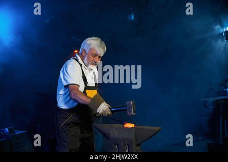 Grauhaariger kaukasischer Handwerker, der mit Industriehammer beheizten Stahl auf Amboss formt. Reifer Schmied, Schürze und Handschuhe, die bei der Schmiede ein Produkt aus geschmolzenem Metall herstellen. Stockfoto