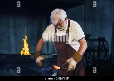 Starker reifer Mann in Schutzhandschuhen und Schürze, die geschmolzenem Stahl mit einer Zepel biegt. Kaukasischer grauhaariger Schmied, der bei der Schmiede Metall auf Amboss formt. Stockfoto