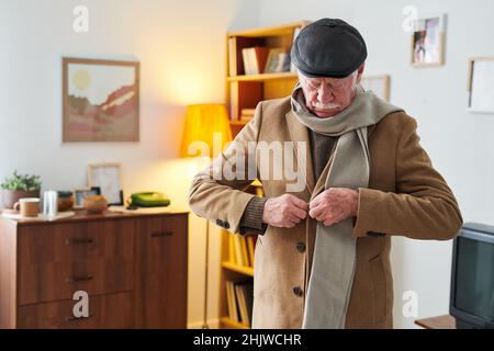 Älterer Mann, der seinen eleganten Mantel für einen Spaziergang anlegt, während er in seiner Wohnung steht Stockfoto