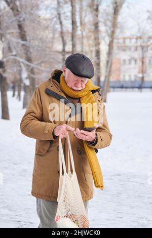 Älterer Mann in warmer Kleidung, der eine Tasche mit einem Lebensmittelgeschäft hält und sein Mobiltelefon benutzt, während er nach dem Einkaufen die Straße entlang läuft Stockfoto