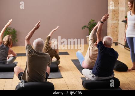 Rückansicht von älteren Menschen, die Dehnungsübungen auf der Trainingsmatte machen, während der Fitnesstrainer sie im Fitnessstudio beobachtet Stockfoto
