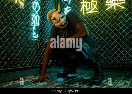 Junger afrikanischer Mann in Maske des Bösen sitzt auf Kniebeugen und berührt den Boden gegen Bars und beleuchtete Hieroglyphen Stockfoto