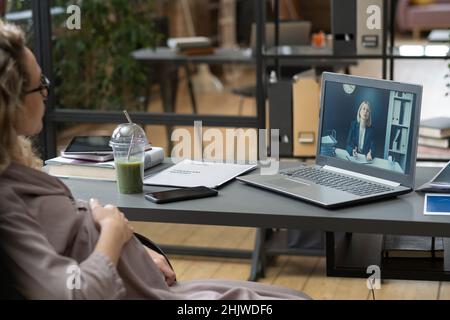 Junge, schwanger arbeitende Büroangestellte kommuniziert mit reifen Kollegen über Arbeitspunkte im Videochat Stockfoto