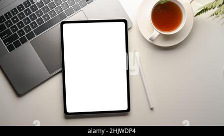 Top-Bild eines modernen Büroschreibtisches mit digitalem Tablet-Computer mit weißem Bildschirm über einem Laptop-Computer auf einem weißen Schreibtisch. Stockfoto