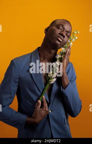 Zufrieden junger afrikanischer Mann in blauer Jacke, der weiße Blumen am Gesicht hält, während er auf gelbem Hintergrund steht Stockfoto