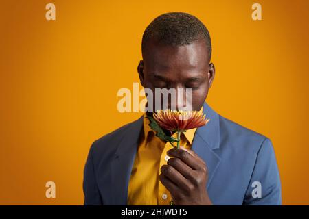 Afroamerikanischer junger Mann in gelbem Hemd und blauer Jacke, der nach Blumen riecht, während er isoliert vor der Kamera steht Stockfoto