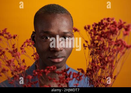 Gesicht eines jungen afrikanischen Mannes, umgeben von roten Blumen, der vor der Kamera auf gelbem Hintergrund steht Stockfoto