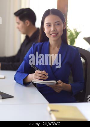 Ein Porträt einer lächelnden asiatischen Geschäftsfrau, die einen Stift und ein Notizbuch am Büroarbeitsplatz hält. Stockfoto