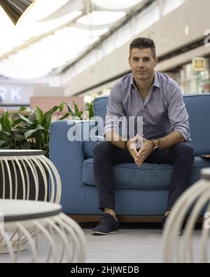 Ein junger, attraktiver Mann, der ein formelles Outfit trägt und auf einer Couch in einem Einkaufszentrum sitzt Stockfoto