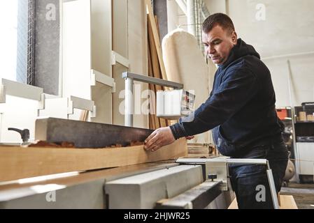 Gelernter Tischler in schwarzer Uniform, der in seiner Holzwerkstätte ein Stück Holz vermessen hat. Horizontale Aufnahme. Selektiver Fokus Stockfoto