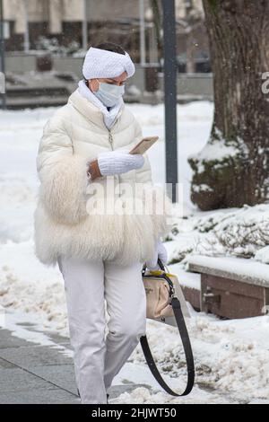 Schönes Mädchen trägt weiße Handschuhe, Fell und Hosen mit schützender medizinischer Maske Überprüfung des Smartphones auf der Straße während Covid oder Coronavirus Stockfoto