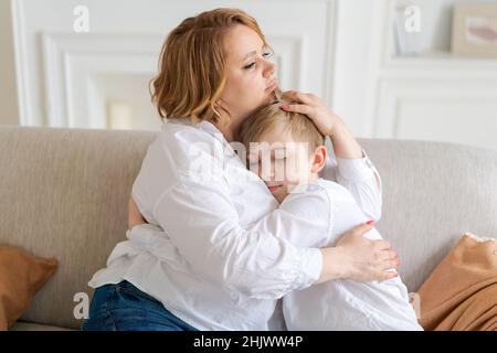 Glücklich niedlich kaukasischen kleinen Sohn umarmt Mutter, während auf dem Sofa im Wohnzimmer zu Hause sitzen. Eine tausendjährige Mutter genießt den Rest mit ihrem Kind, umarmt das Kind. Mutterschaft Konzept Stockfoto