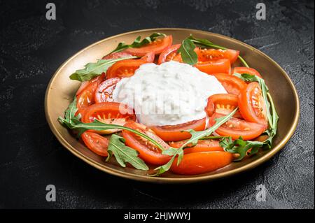 Italienische Salatkaprese mit Käse Stracciatella, Tomaten und Rucola. Schwarzer Hintergrund. Draufsicht Stockfoto