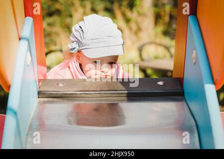 Unentschlossenheit blondes Haar Kleinkind Mädchen in lustigen Hut auf einer Rutschung auf einem Spielplatz in einem öffentlichen Park Stockfoto