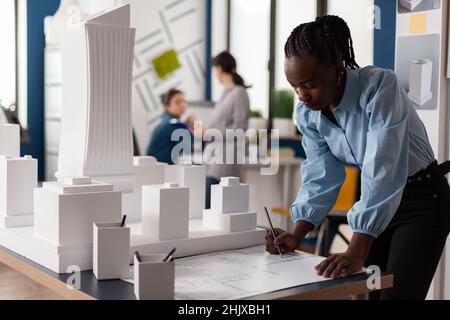 Wohnarchitekt, der am Schreibtisch im Architekturbüro neben einem Wolkenkratzer im weißen Schaumstoffmaßstab Baupläne zeichnet. Entwicklungsingenieur macht sich Notizen zum Bauplan. Stockfoto