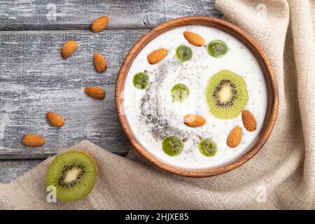 Joghurt mit Kiwi, Stachelbeere, Chia und Mandeln in Holzschüssel auf grauem Holzhintergrund und Leinentextilien. Draufsicht, flaches Lay, Nahaufnahme. Stockfoto