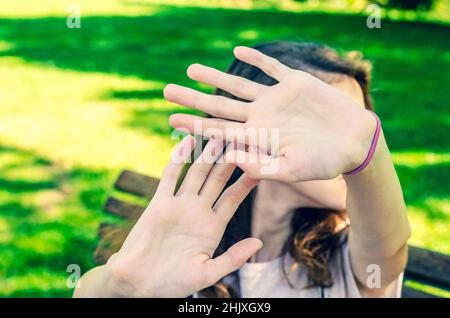 Schönes junges Mädchen, das in einem Park sitzt und das Gesicht mit den Händen versteckt Stockfoto